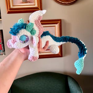 May include: A handmade crochet dragon in white, pink, blue, and teal. The plush toy has large, iridescent eyes and a long, curved tail. The dragon's head features a tuft of blue fur and small, green accents. The dragon is held in a person's hand.