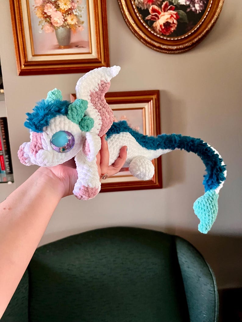 May include: A handmade crochet dragon in white, pink, blue, and teal. The plush toy has large, iridescent eyes and a long, curved tail. The dragon's head features a tuft of blue fur and small, green accents. The dragon is held in a person's hand.