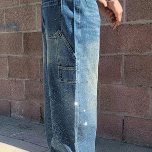 May include: A pair of blue denim carpenter jeans with rhinestones. The jeans have a relaxed fit and are cuffed at the ankles.