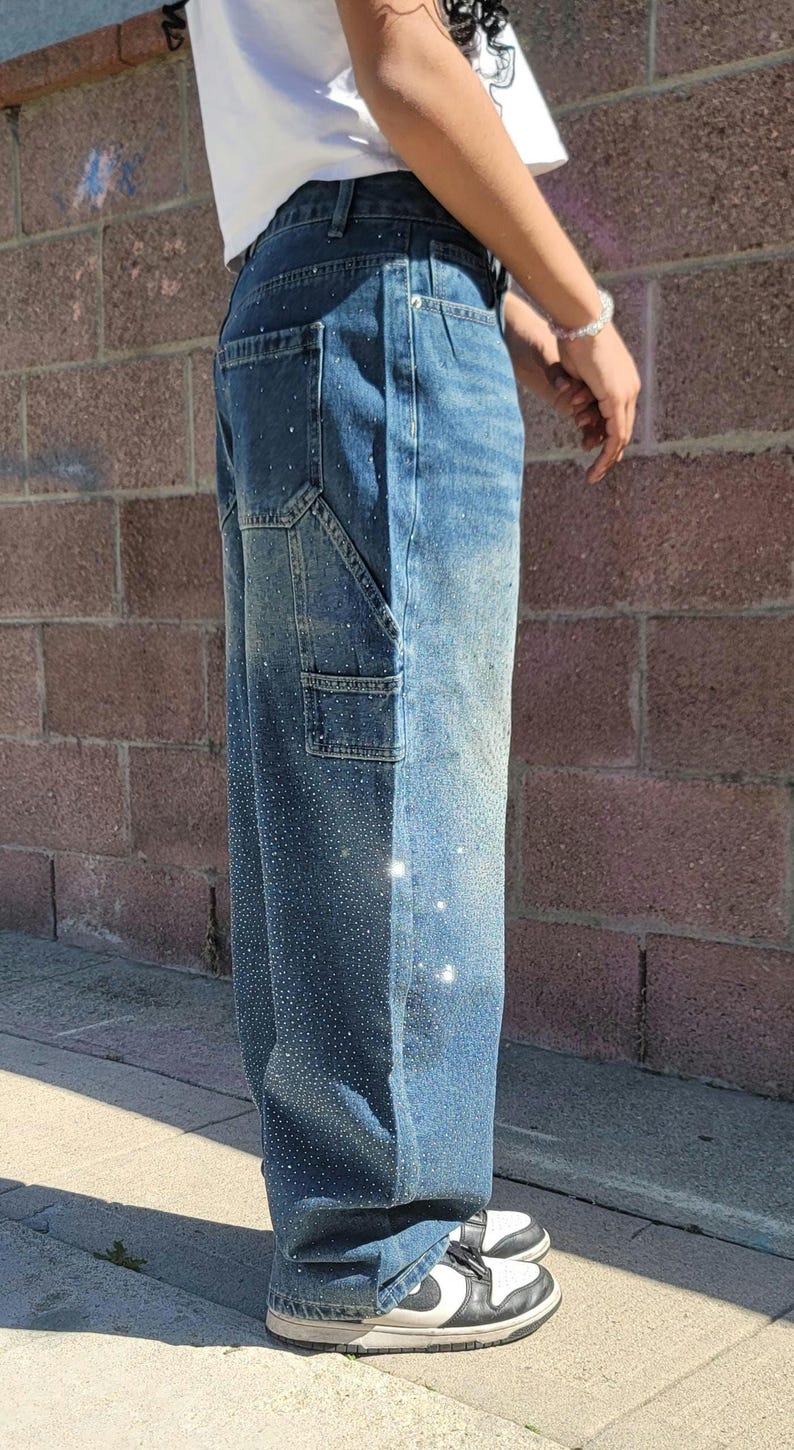 May include: A pair of blue denim carpenter jeans with rhinestones. The jeans have a relaxed fit and are cuffed at the ankles.