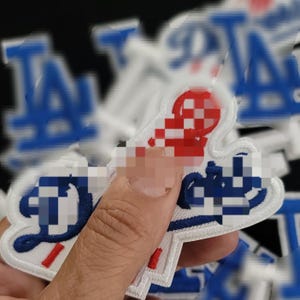Peut inclure: Écusson brodé avec le logo des Dodgers de Los Angeles en bleu, blanc et rouge. Le patch a une bordure blanche et est tenu par un doigt. L'arrière-plan est flou avec d'autres écussons.