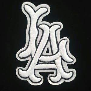 May include: White embroidered patch with the letters "LA" in a decorative script, edged with black. The patch is on a black background, creating a strong contrast. Ideal for customising clothing or accessories. The patch is approximately 7 cm in height.