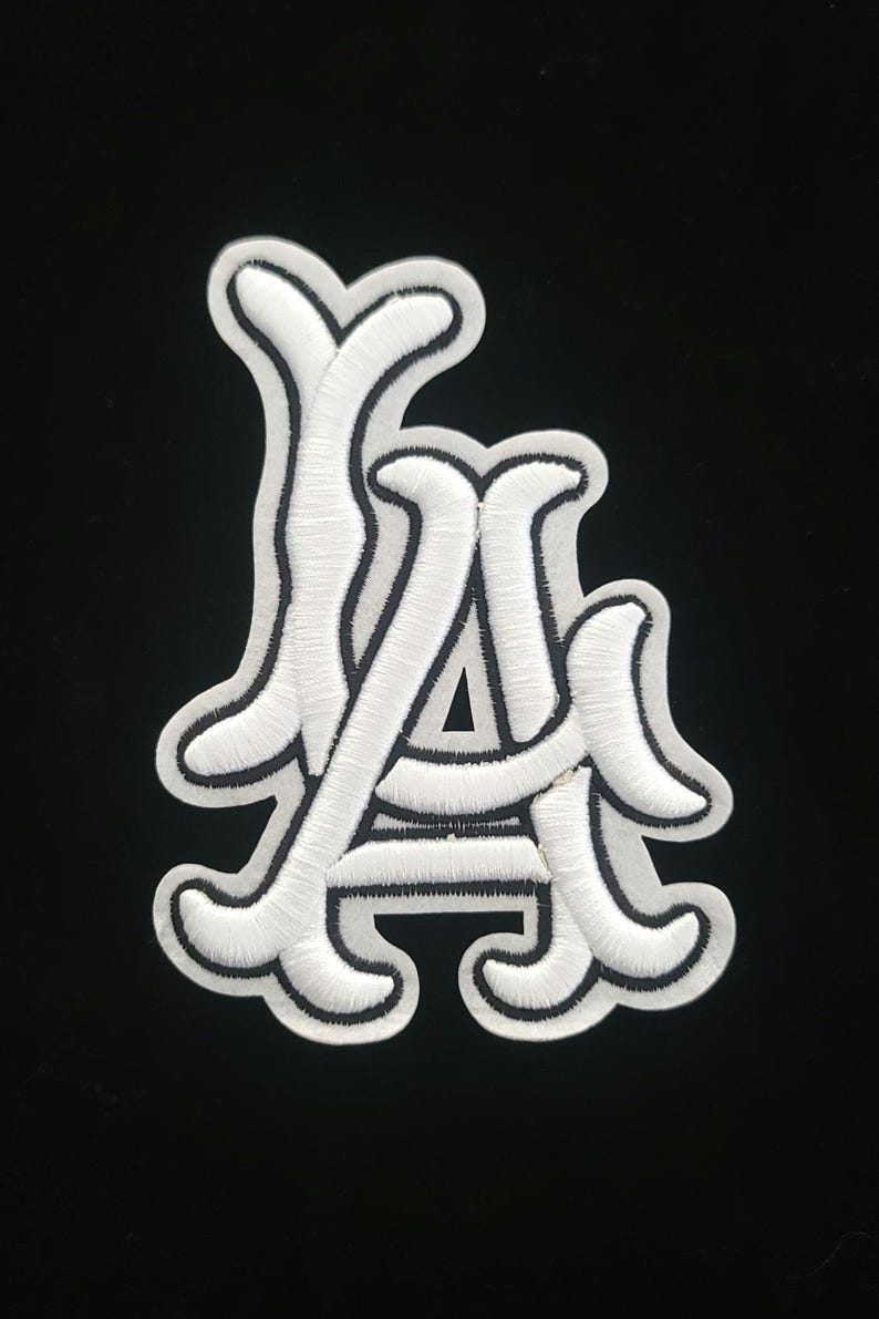 May include: White embroidered patch with the letters "LA" in a decorative script, edged with black. The patch is on a black background, creating a strong contrast. Ideal for customising clothing or accessories. The patch is approximately 7 cm in height.