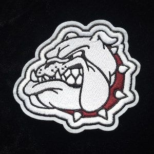 May include: Embroidered patch featuring a white bulldog head with a fierce expression. The bulldog has a black outline and details, with a red spiked collar. The patch has a white border and is set against a black background.