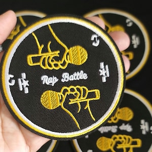 May include: Black round patch with a yellow and white border. The patch features a yellow illustration of a hand holding a microphone, with the words "Rap Battle" in white. Additional patches are visible in the background.