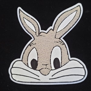 May include: A Bugs Bunny patch with a white outline. The patch features a detailed image of Bugs Bunny's face, with large eyes, long ears, and a textured, light brown fur appearance. The background is black.