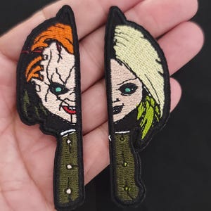 May include: Two embroidered patches in the shape of knives, each featuring a character's face. One has orange hair and a scarred face, the other has blonde hair. The knife handles are olive green.