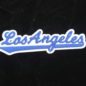 Pu&ograve; includere: Toppa ricamata con la scritta "Los Angeles" in blu, in corsivo e bordata di bianco. Il testo &egrave; posto su uno sfondo nero. Ideale per personalizzare abbigliamento o accessori.