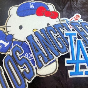 May include: An embroidered patch featuring a white cartoon cat wearing a blue baseball cap with the letters LA, a red bow, and a blue glove. The patch includes the text LOS ANGELES in blue block letters with black outlines and a blue LA logo.