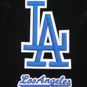 May include: Embroidered patch featuring the Los Angeles Dodgers logo, a stylized "LA" in blue with a white border, and the words "Los Angeles" in script below. The patch is set against a black background.