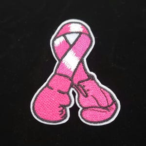 May include: Embroidered patch featuring a pink ribbon and pink boxing gloves. The ribbon has a white stripe in the center. The patch has a white border and is set against a black background. The design is a symbol of support.