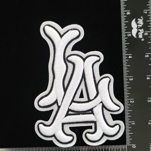 May include: White embroidered patch featuring the letters "LA" in a stylized script with a black border. The patch is on a black background. The patch is approximately 7.6 cm wide.