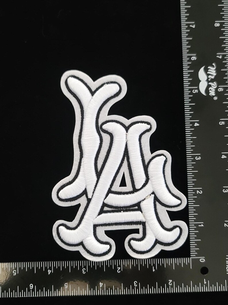 May include: White embroidered patch featuring the letters "LA" in a stylized script with a black border. The patch is on a black background. The patch is approximately 7.6 cm wide.