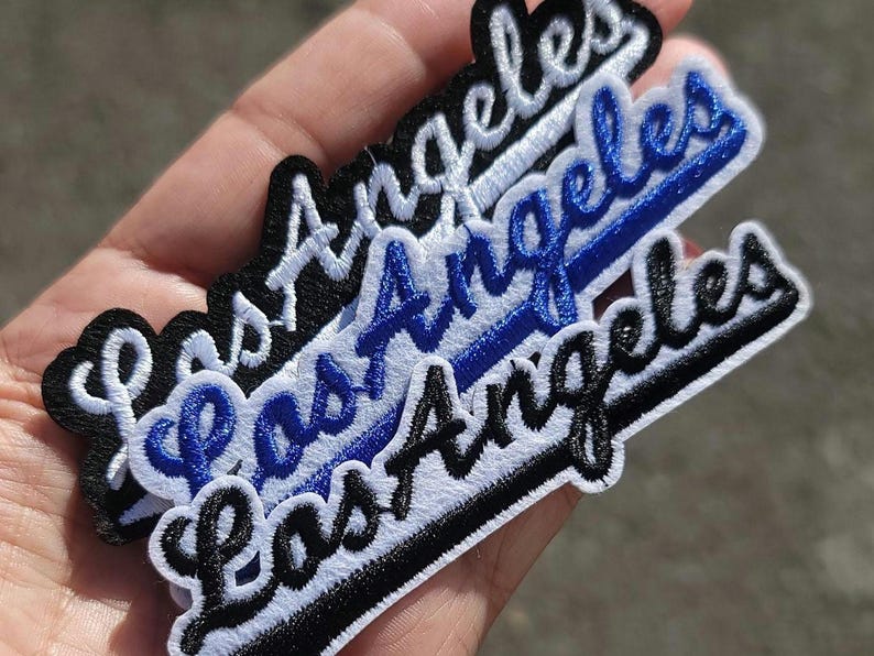 Pu&ograve; includere: Toppe ricamate con la scritta "Los Angeles" in corsivo. Le toppe sono in nero, bianco e blu, con un bordo bianco. Le toppe sono impilate l'una sull'altra.
