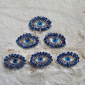 May include: Six blue and gold evil eye charms with white backgrounds. The charms are made of rhinestones and have a blue eye with a gold pupil and a gold outline.