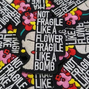 Peut inclure: Écusson brodé avec un portrait stylisé d'une femme orné de fleurs. Le texte "NOT FRAGILE LIKE A FLOWER FRAGILE LIKE A BOMB" est en blanc sur fond noir. Il y a des détails floraux rouges, roses et jaunes.