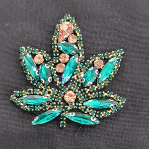 May include: A decorative leaf-shaped embellishment, featuring a cannabis leaf design. The leaf is adorned with emerald green and peach-colored rhinestones, outlined with gold-colored metal. The embellishment is set against a black fabric background.