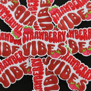 May include: Embroidered patches with the words "STRAWBERRY VIBES" in red, bubble-style letters. Each patch features a strawberry graphic with green leaves and pink fruit. The patches are on a white background.
