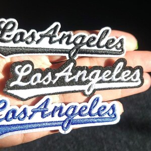 Pu&ograve; includere: Tre toppe ricamate con la scritta "Los Angeles" in un carattere corsivo. Le toppe sono in bianco e nero e in blu e bianco. Le toppe sono probabilmente per abbigliamento o accessori.