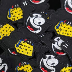 Peut inclure: Plusieurs écussons brodés noirs représentant un ours de dessin animé souriant avec un grand sourire et un bloc de fromage jaune. L'ours a des yeux blancs et une bouche rouge. Le bloc de fromage a des détails bleus et jaunes.