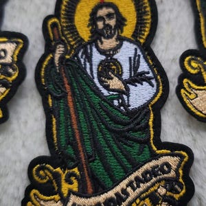 May include: Embroidered patch featuring a saint with a halo, holding a staff and wearing a green robe. The patch has a gold background and the text "San Judas Tadeo" on a banner.