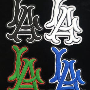 May include: Four embroidered patches displaying the letters "LA" in a stylized design. The patches are in black with white trim, white with black trim, green with red trim, and blue with white trim. The background is black.