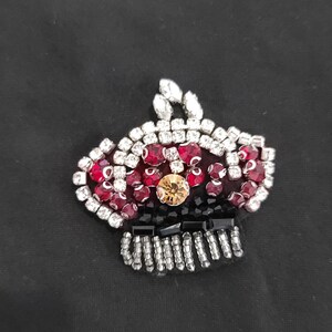 May include: A jewelled crown brooch on a black background. The crown is adorned with red and clear rhinestones, a central gold gem, and black and silver bead details. Suitable for clothing or accessories.