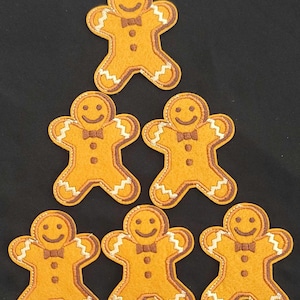 May include: Six gingerbread man patches arranged on a black background. Each patch is a smiling gingerbread man with a brown bow tie, brown outline, and white zig-zag trim. The gingerbread men are a warm, golden-brown color.