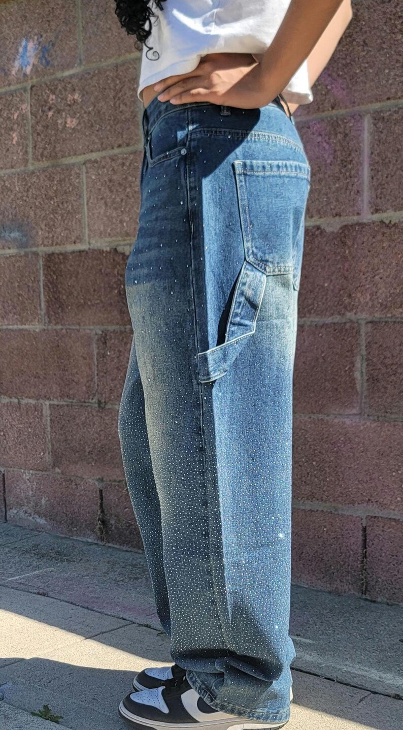 May include: A pair of blue denim carpenter jeans with rhinestones. The jeans have a relaxed fit and a wide leg.