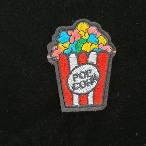 May include: A red and white striped popcorn bucket with a white circle containing the text "POP CORN" in black. The bucket is filled with colorful popcorn, including yellow, pink, blue, and green.