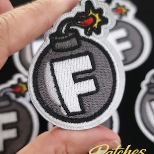 May include: Embroidered patch featuring a gray bomb design with a white "F" in the center. The bomb has a black outline and a red and yellow explosion. The patch is on a black background.