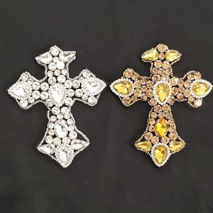 May include: Two decorative cross-shaped embellishments. One is silver-toned with clear crystals, the other is gold-toned with amber and yellow crystals. Both have a detailed, ornate design and are set against a black background.