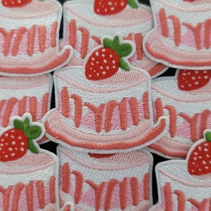 Peut inclure: Écussons brodés représentant un gâteau rose et blanc avec une fraise sur le dessus. Les écussons ont une bordure rose et le mot "YUM" est brodé sur le gâteau. Les écussons sont de couleur rose clair et blanc.
