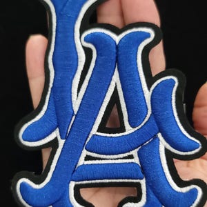 May include: Embroidered patch with the letters "LA". The letters are in blue with white and black edging. The patch is designed for application on clothing or accessories.