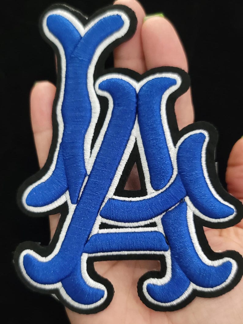 May include: Embroidered patch with the letters "LA". The letters are in blue with white and black edging. The patch is designed for application on clothing or accessories.