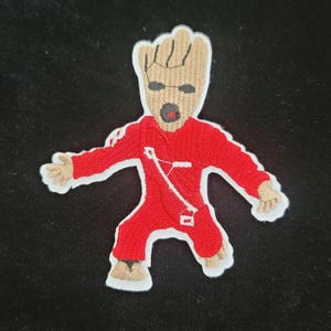 May include: An embroidered patch depicting a fictional character with a brown head, black eyes, and a red mouth. The character is wearing a red jumpsuit with white accents.
