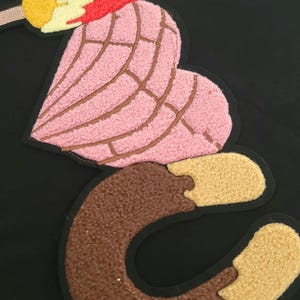 May include: An embroidered patch with a pink concha pastry design, brown lines, a brown letter "C", and a yellow and red popsicle. The patch is set against a black background.