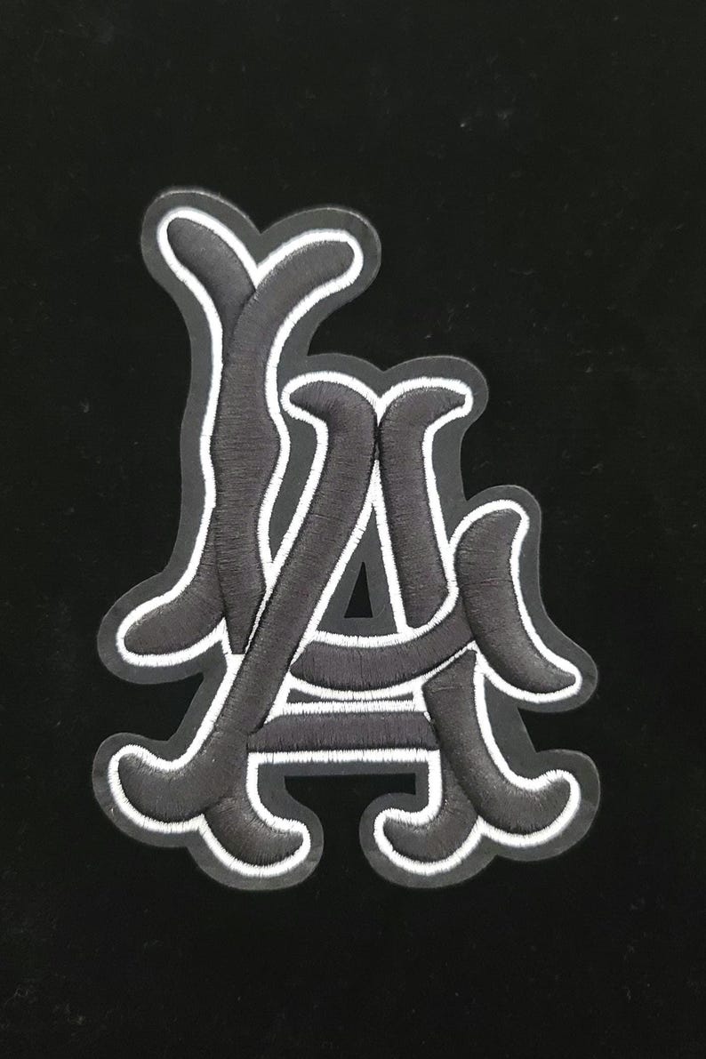May include: Embroidered patch in black and white, displaying the letters "LA" in a decorative font. The black letters have a white border, set against a black backdrop. Ideal for customising clothing or accessories.