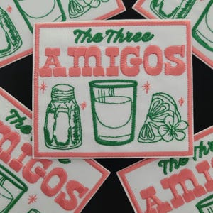 May include: Embroidered patch with the text "The Three AMIGOS" in pink and green. The design includes a salt shaker, a shot glass, and a lime wedge. The patch has a white background and a pink border.