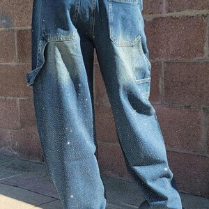 May include: A pair of blue denim jeans with a loose fit and a rhinestone design. The jeans have a relaxed, baggy silhouette and feature a subtle distressed look.