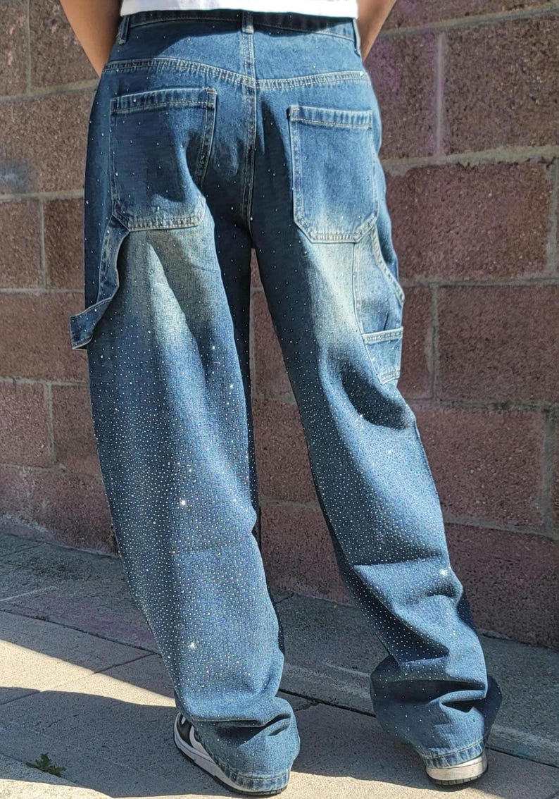 May include: A pair of blue denim jeans with a loose fit and a rhinestone design. The jeans have a relaxed, baggy silhouette and feature a subtle distressed look.