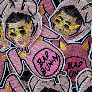 May include: Pink embroidered patches featuring a cartoon image of a person wearing a bunny costume and yellow sunglasses. The patches have the words "Bad Bunny" in a pink oval.