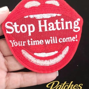 May include: A red embroidered patch shaped like an open mouth with white teeth. The text Stop Hating is embroidered in white across the upper teeth, and the text Your time will come! is embroidered in white across the lower lip area.