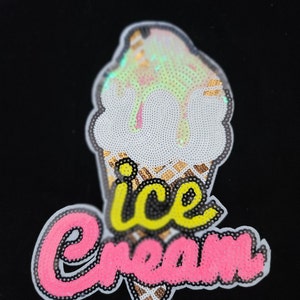 Puede incluir: Un cono de helado blanco y dorado con un texto "helado" rosa y amarillo. El cono de helado está hecho de lentejuelas.