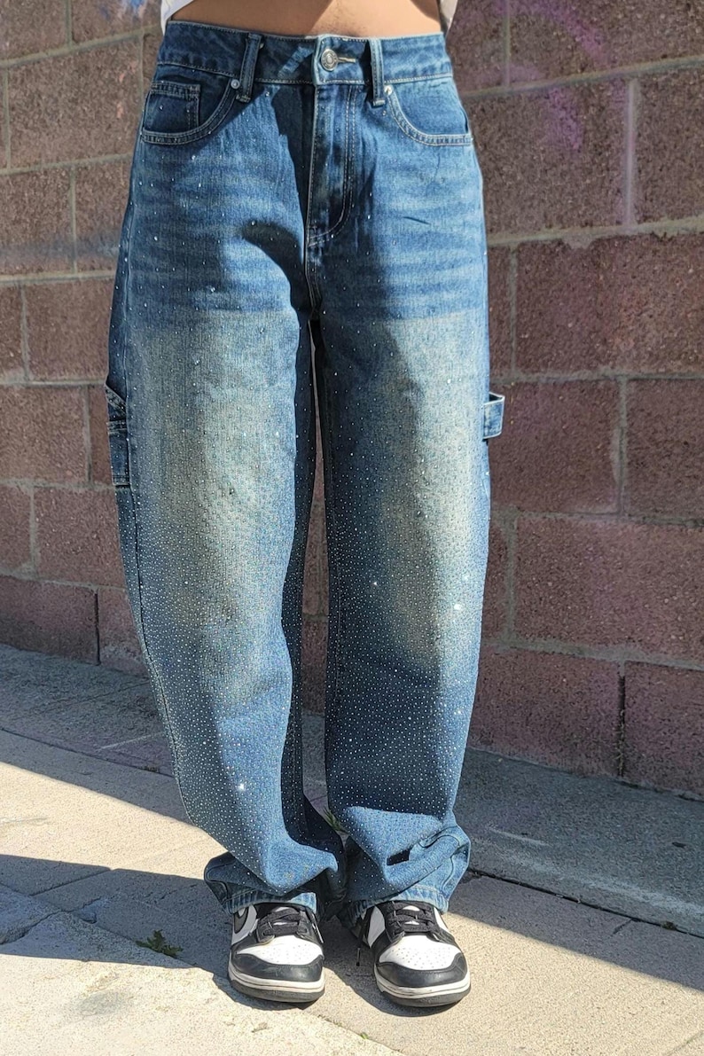 May include: A pair of blue denim carpenter jeans with rhinestones. The jeans have a relaxed fit and are worn with black and white sneakers.