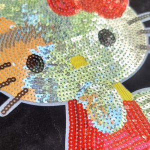 May include: A decorative patch featuring a stylized cat face crafted from shimmering sequins in shades of pale yellow, orange, and red. The design includes a bright red bow, dark circular eyes, and a small yellow nose, all set against a black background.