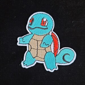 May include: Embroidered patch of a Squirtle cartoon character. The patch has a light blue body, a tan shell, and red details. The character is outlined in white and set against a black background.