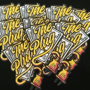 May include: A collection of embroidered patches with the words "The Plug" in yellow script. The patches feature a design of a stack of money and a plug. The patches are yellow, red, white, and black.