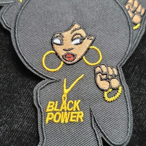 May include: Black and gold embroidered patch featuring a cartoon woman with an afro, large hoop earrings, and a raised fist. The text "BLACK POWER" is embroidered in gold below the woman.