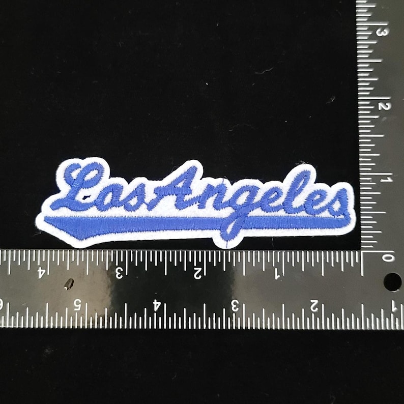Pu&ograve; includere: Toppa ricamata blu e bianca con la scritta "Los Angeles" in corsivo. La toppa ha un bordo bianco e un contorno blu, con una barra blu sotto il testo. Misura circa 10 cm di lunghezza.